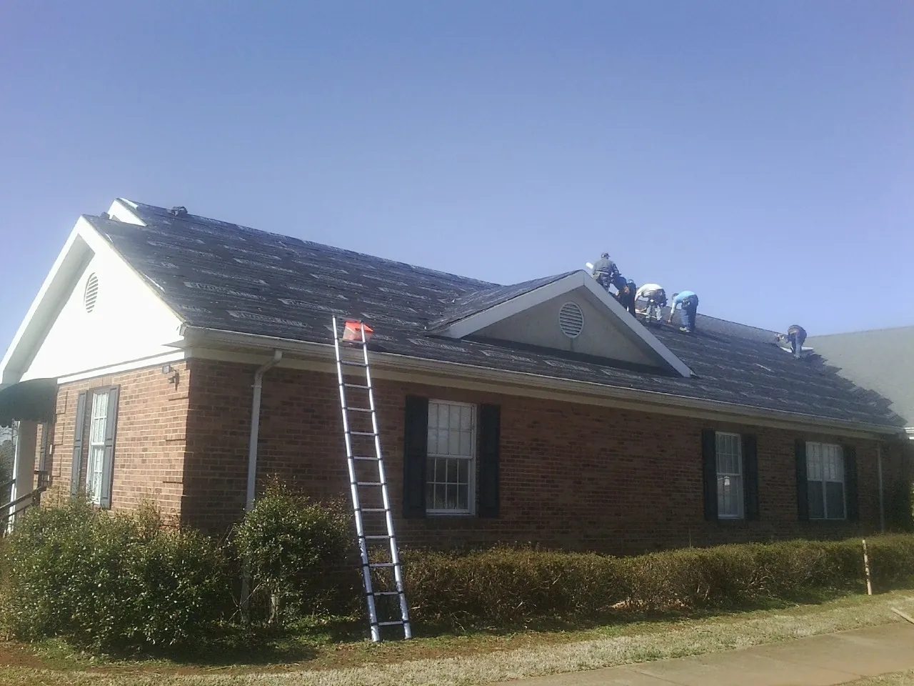 Crew tearing shingles off a brick ranch roof with a ladder set — Emergency Roof Repair work by our crew in Ixonia