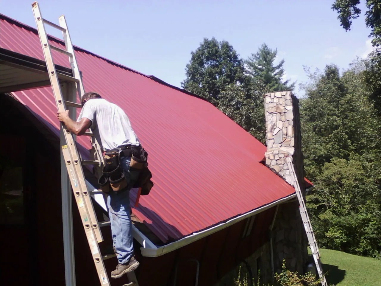 Roofer on a ladder beside a red metal roof and stone chimney — Soffit & Fascia Repair by Roofing Brothers in Ixonia